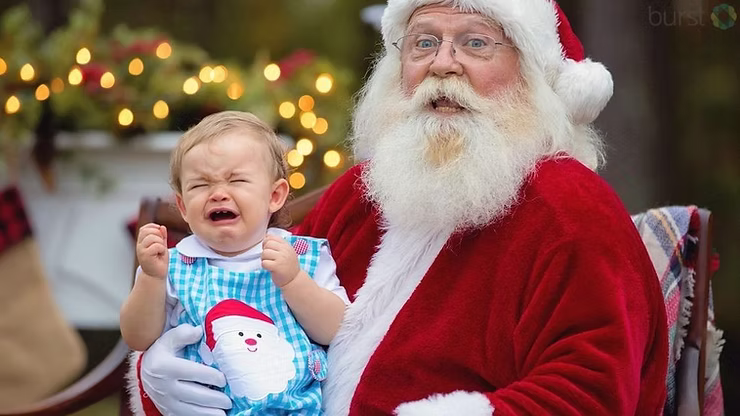 A baby in a blue checkered outfit with a Santa Claus graphic is crying while being held by a man dressed as Santa Claus, with blurred Christmas lights in the background.