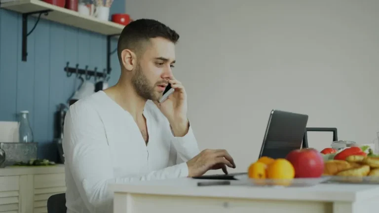 A man in a white shirt is talking on his phone and typing on a laptop in a kitchen. A fruit bowl with apples and lemons is on the counter