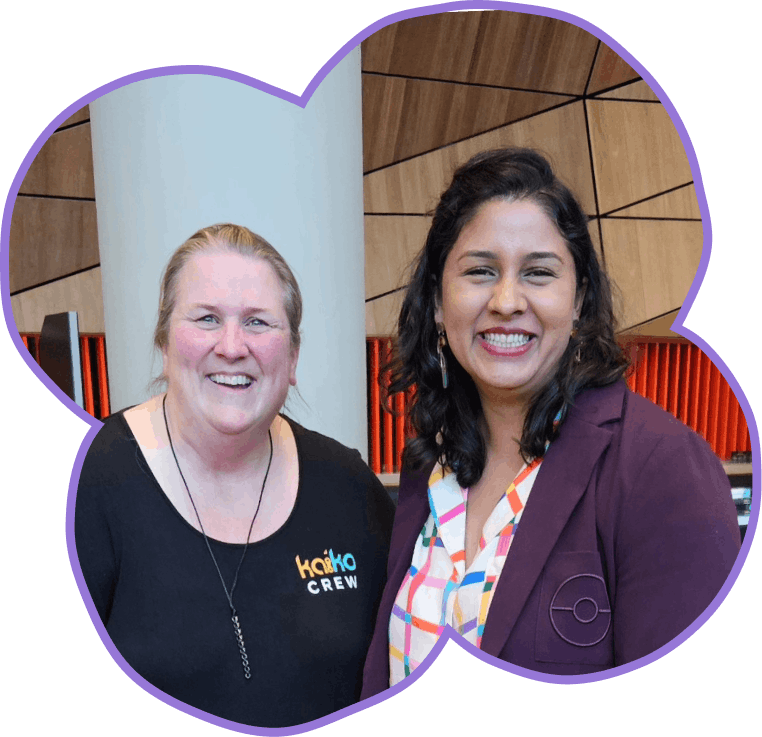 Two women smile at the camera. The woman on the left wears a black t-shirt with "kaiko CREW" printed on it. The woman on the right wears a purple blazer over a colorful patterned shirt