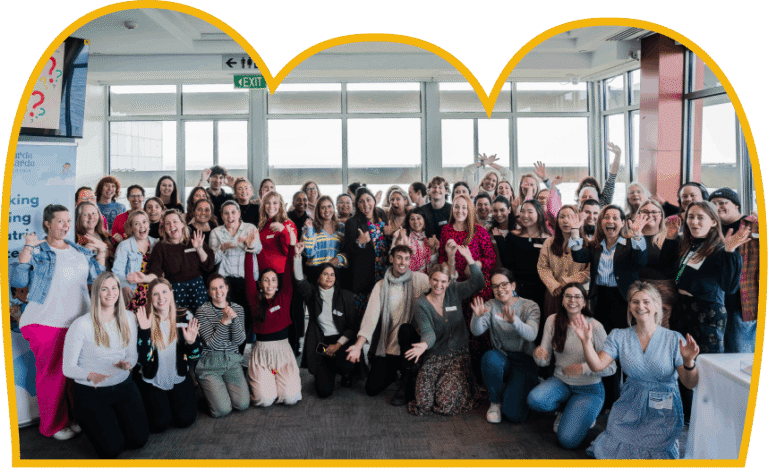 A large group of smiling women pose for a photo, many with hands raised. They are gathered indoors, in front of a wall of windows and a banner