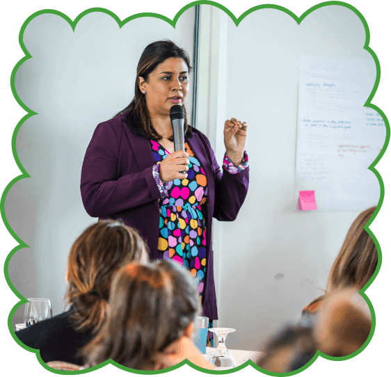 A woman in a colorful polka dot dress and purple blazer speaks into a microphone at a workshop, gesturing with her free hand while a whiteboard with notes is visible behind her