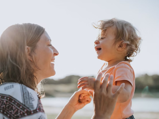 A smiling adult woman and a laughing toddler are looking at each other outdoors with the sun shining behind them.