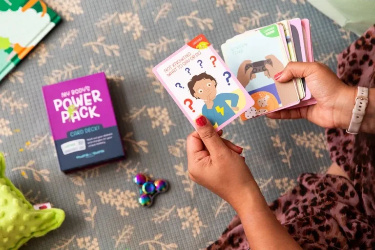 A person's hands hold a stack of colorful cards, with one card showing an illustration of a child and question marks. A purple box labeled "My Body's Power Pack" sits nearby on a patterned rug.